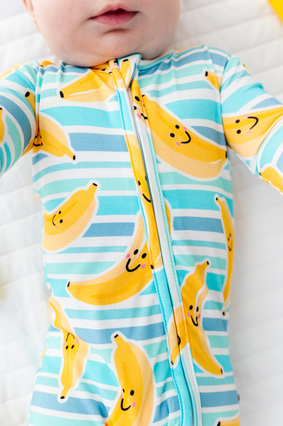A close-up of a baby wearing a long-sleeve zip-front footed sleeper with a light blue and white striped pattern and yellow banana print with smiling faces.