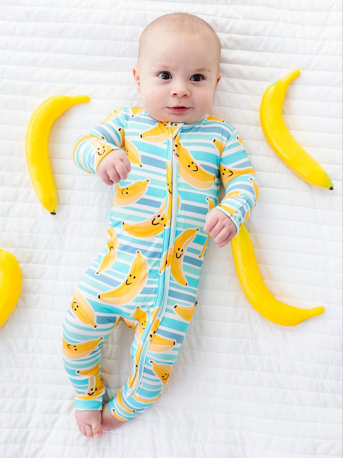 A baby lying on a white quilted blanket wearing a one-piece zip-up sleeper with a yellow banana print and light blue stripes. Yellow toy bananas are placed around the baby.