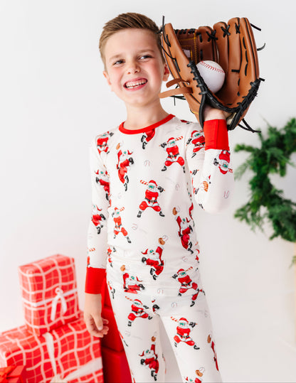 A young boy smiling while wearing bamboo Christmas-themed Santa Claus pajamas, holding a baseball glove with a baseball inside, with a green wreath in the background.