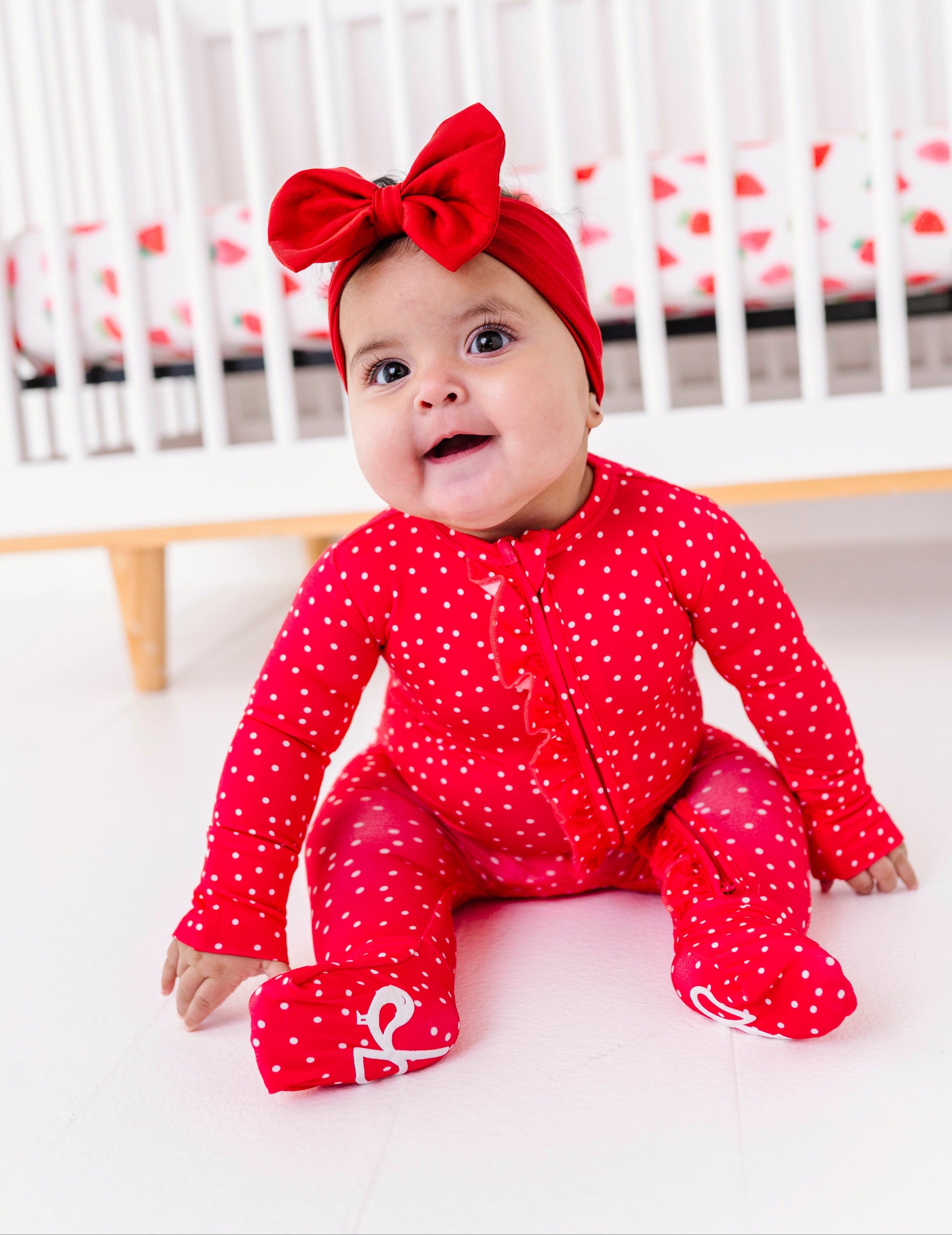 Baby in a red romper with a bow sitting in front of a crib.