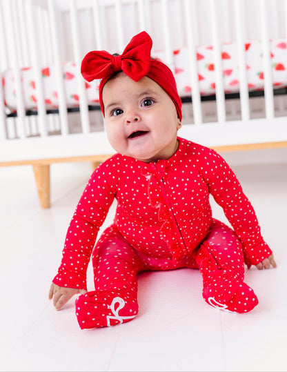 Baby in a red romper with a bow sitting in front of a crib.