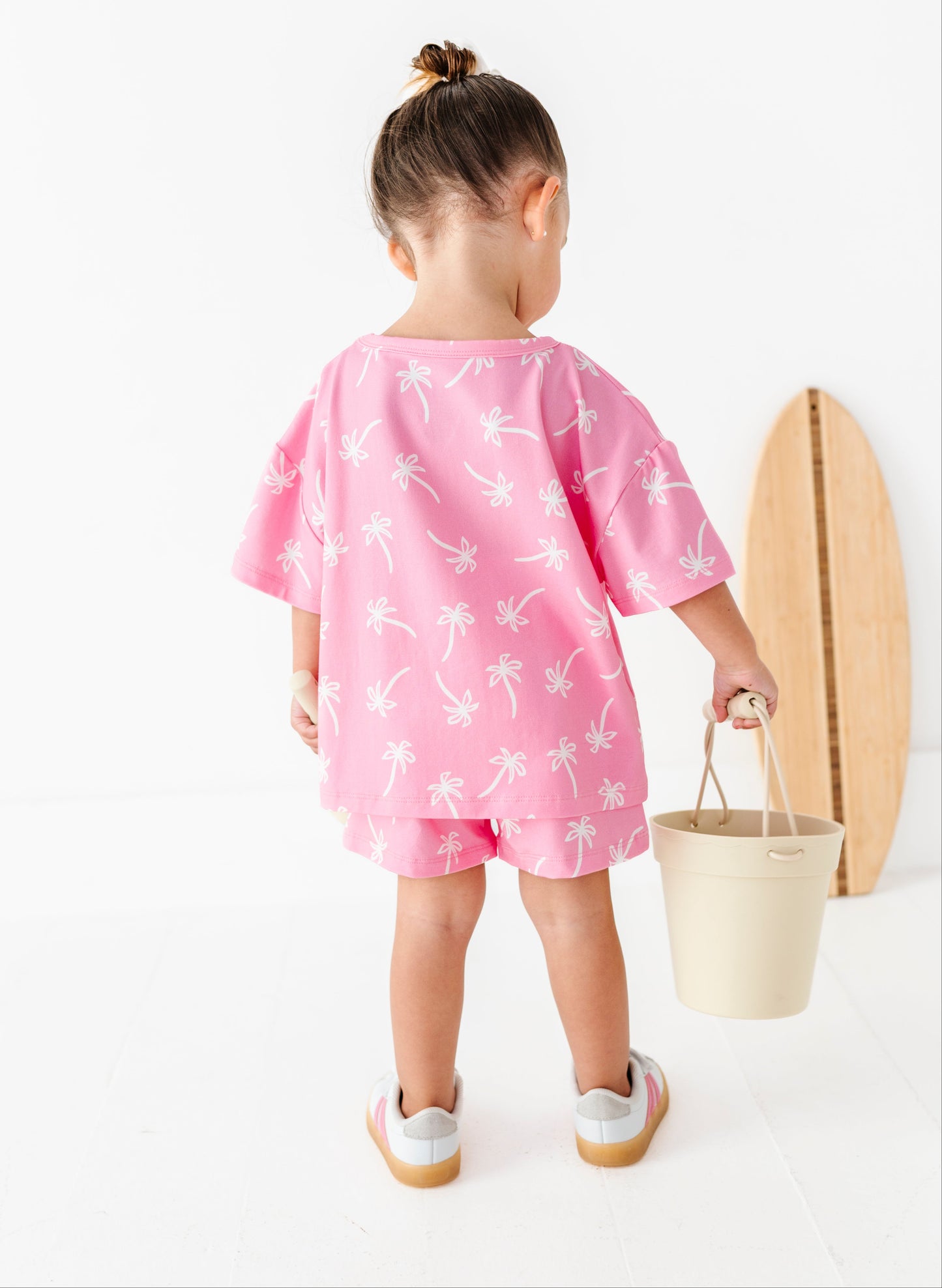 Child wearing a pink dress with white patterns, holding a bucket and wooden surfboard on a white background