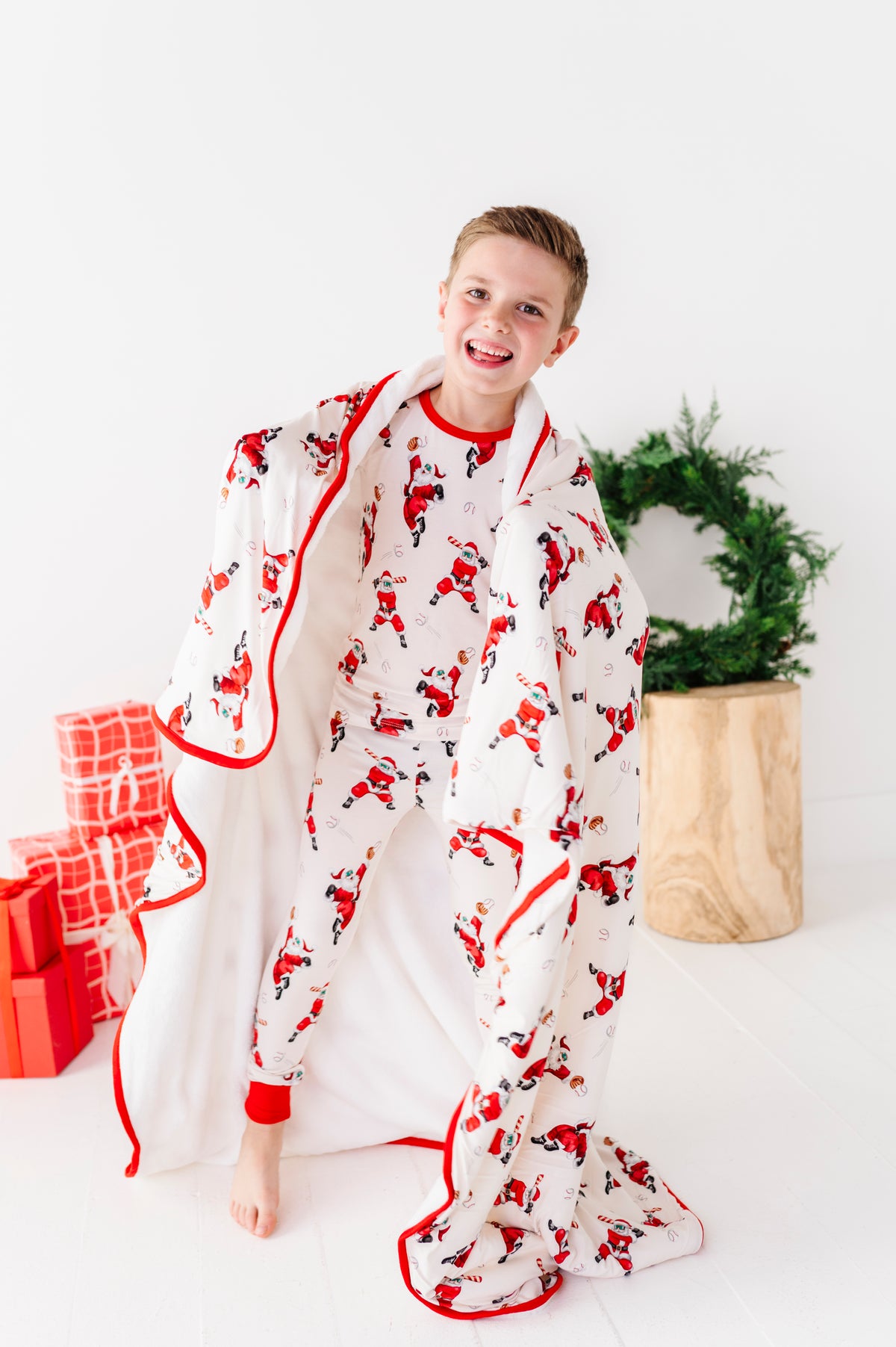 A young boy wearing bamboo pajamas featuring a variety of Santas playing baseball. He's wrapped in a matching baseball Santa bamboo blanket and standing in front of a green wreath. 