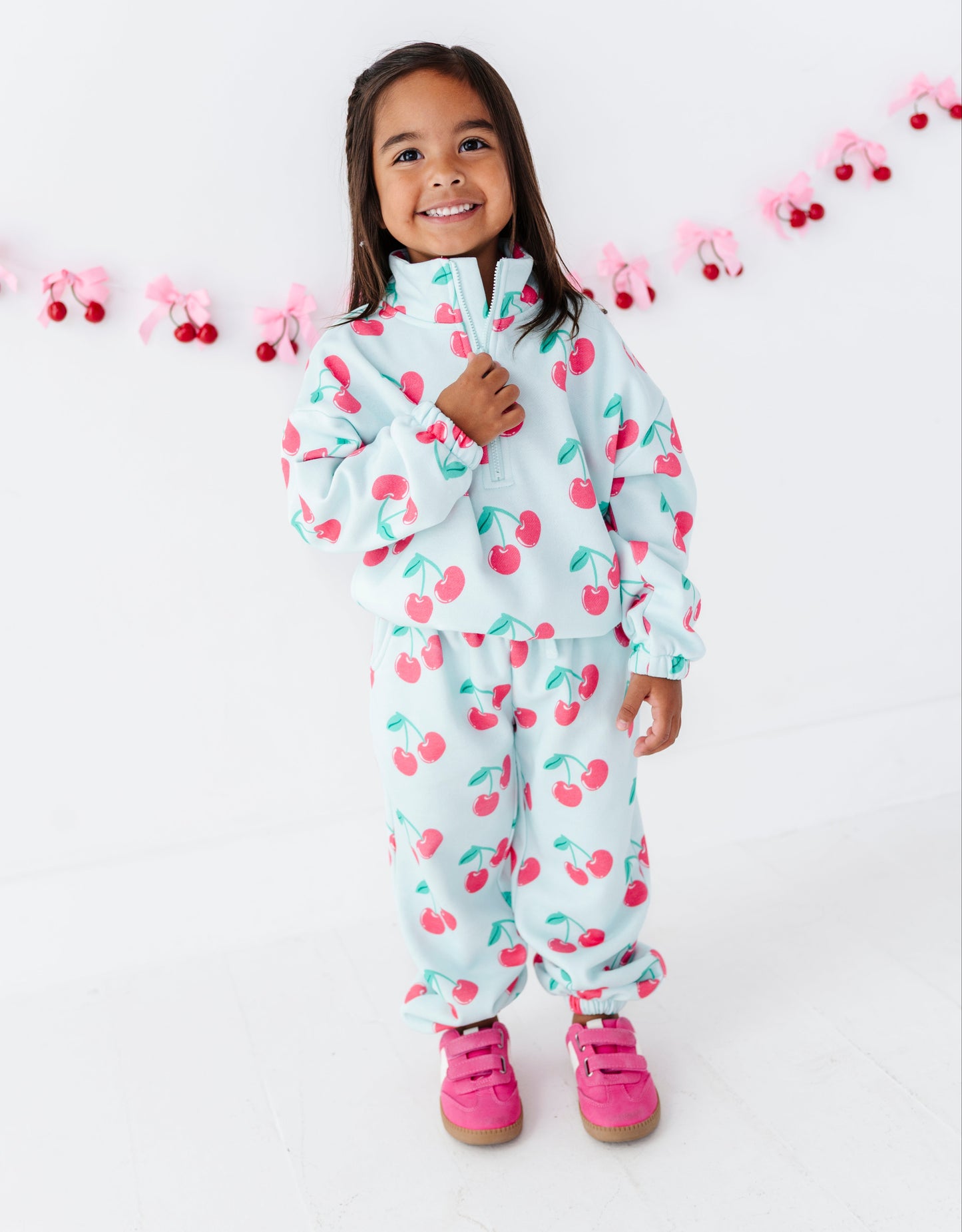 A young girl smiling while wearing a matching white two-piece tracksuit with a pink and green cherry print, paired with pink sneakers, set against a white background with a pink garland.