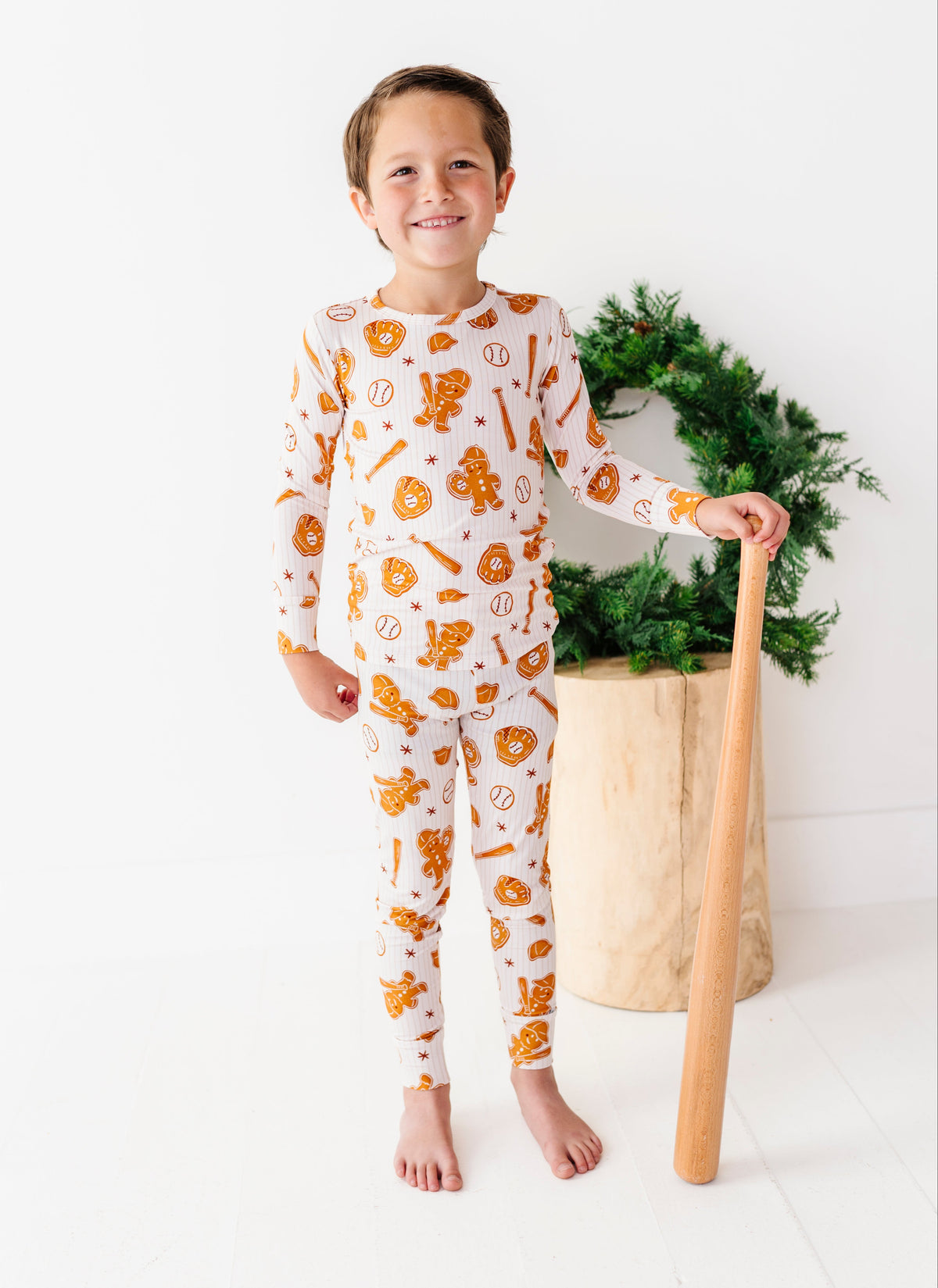 A young boy stands barefoot in a bright white room, wearing a long-sleeve pajama set with an orange and yellow pumpkin and cookie pattern. He is holding a wooden toy bat next to a green wreath in a wooden pot.