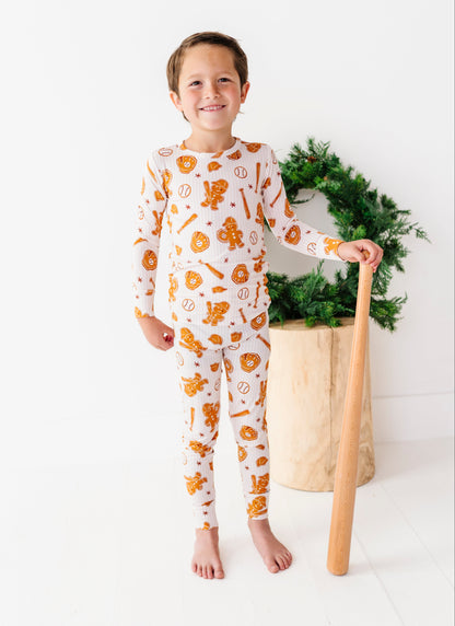 A young boy stands barefoot in a bright white room, wearing a long-sleeve pajama set with an orange and yellow pumpkin and cookie pattern. He is holding a wooden toy bat next to a green wreath in a wooden pot.
