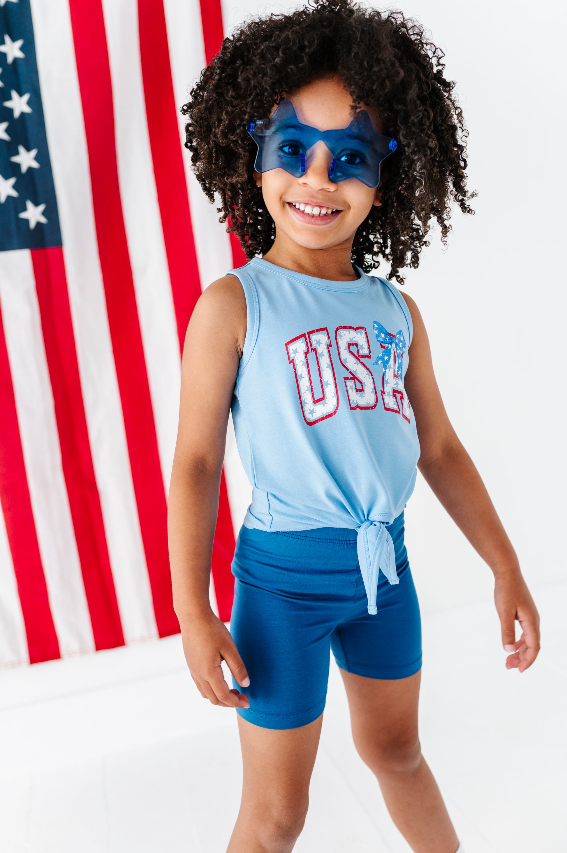 Child wearing a Fourth of July tank top, paired with bamboo biker shorts, standing in front of an American flag.
