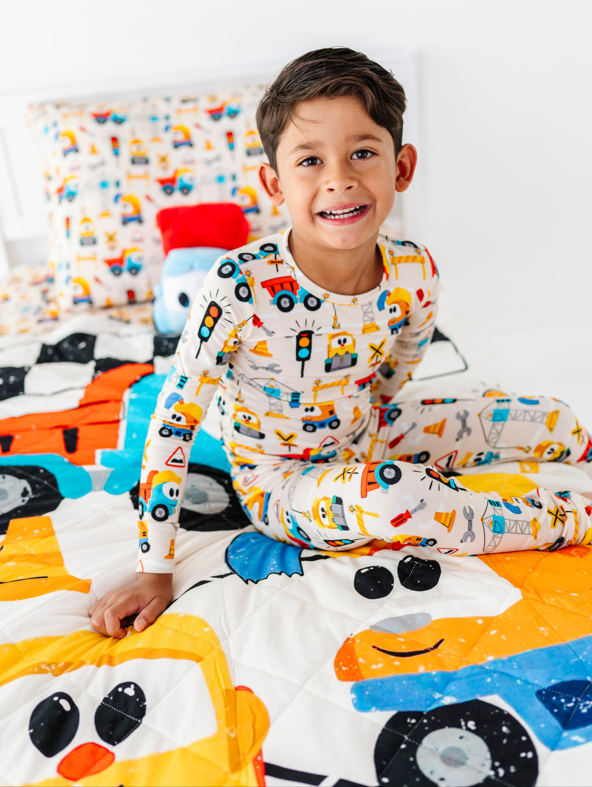 A smiling young boy wearing a white long-sleeve pajama set with a colorful construction vehicle print, sitting on a bed with a matching colorful truck-themed quilt.