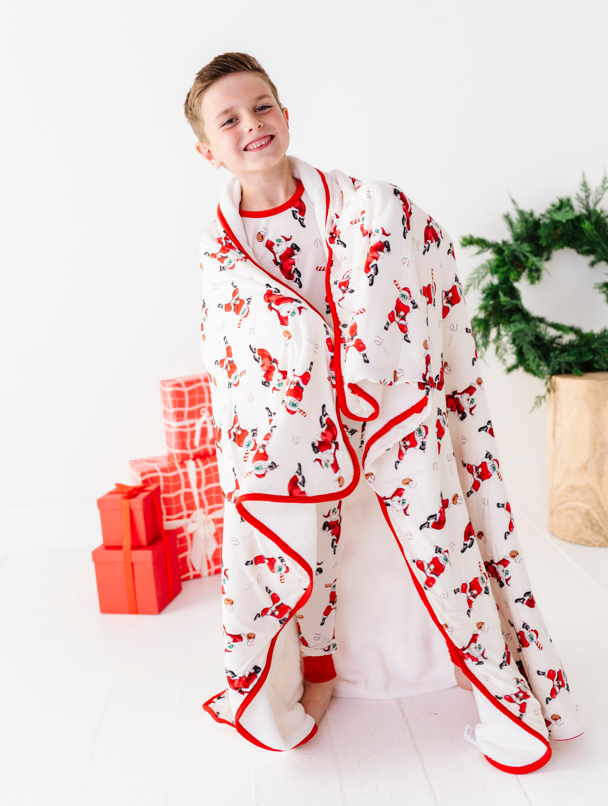 A smiling young boy wearing a white Santa Claus print pajama robe with red trim, standing in a bright white room with wrapped gifts and a green wreath in the background.