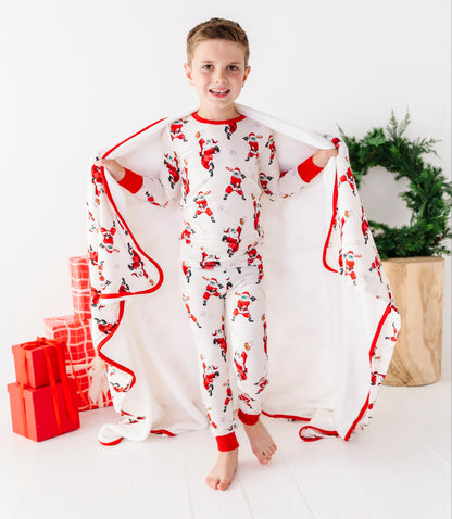 A young boy stands barefoot in a bright white room, wearing a white Santa Claus pajama set and holding open a matching white blanket with red trim. Red wrapped gift boxes sit to his left, and a green wreath in a wooden planter is to his right.
