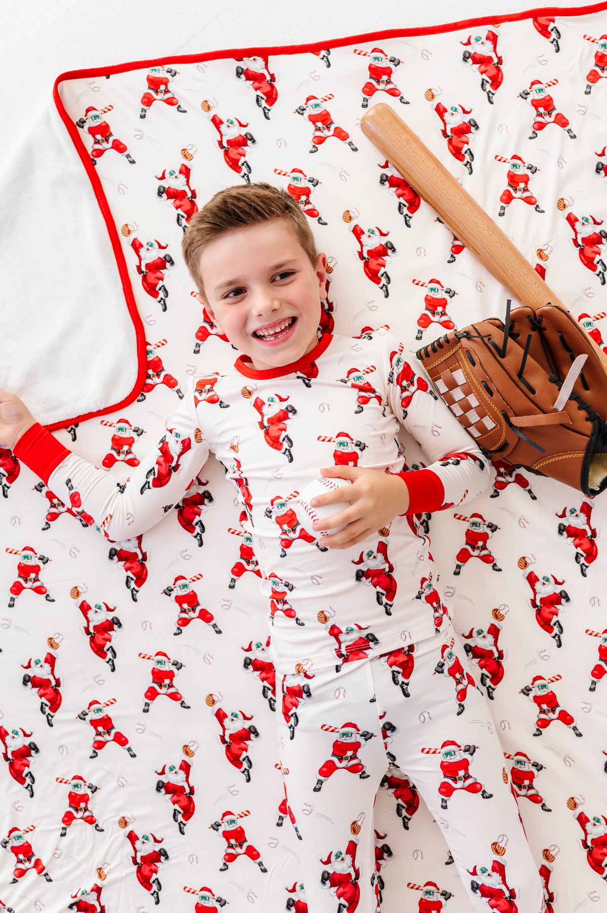 A young boy wearing a long-sleeve bamboo pajama top and matching pants with an all-over Santa Claus baseball print, laying on a matching baseball Santa bamboo blanket with his wooden baseball bat and leather glove. 