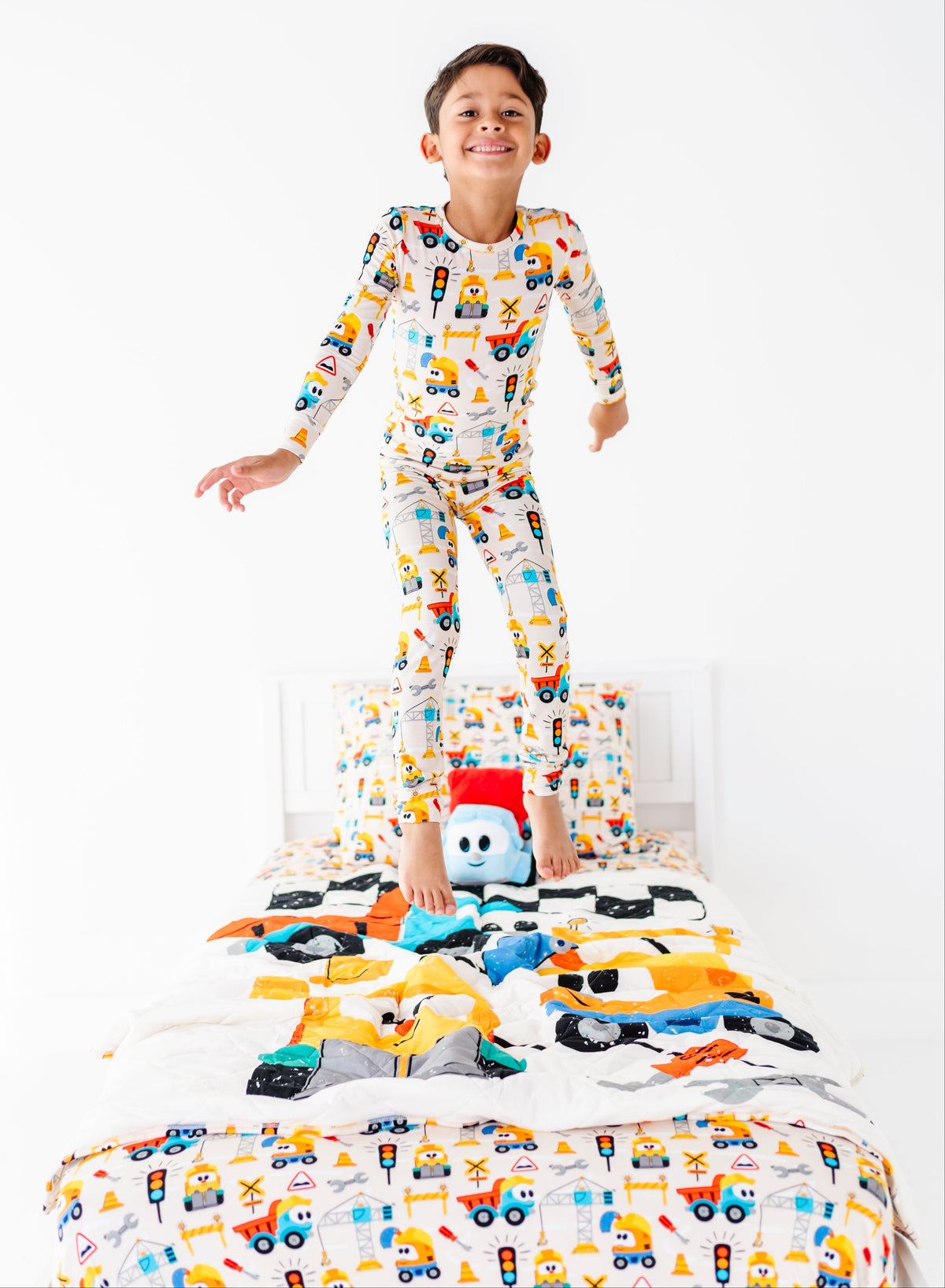 A young boy stands on a bed wearing a long-sleeve pajama set with a colorful construction vehicle print. The room has a clean white background, and a small plush toy is on the bed in front of him.