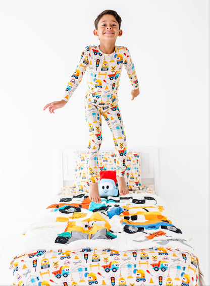 A young boy stands on a bed wearing a long-sleeve pajama set with a colorful construction vehicle print. The room has a clean white background, and a small plush toy is on the bed in front of him.