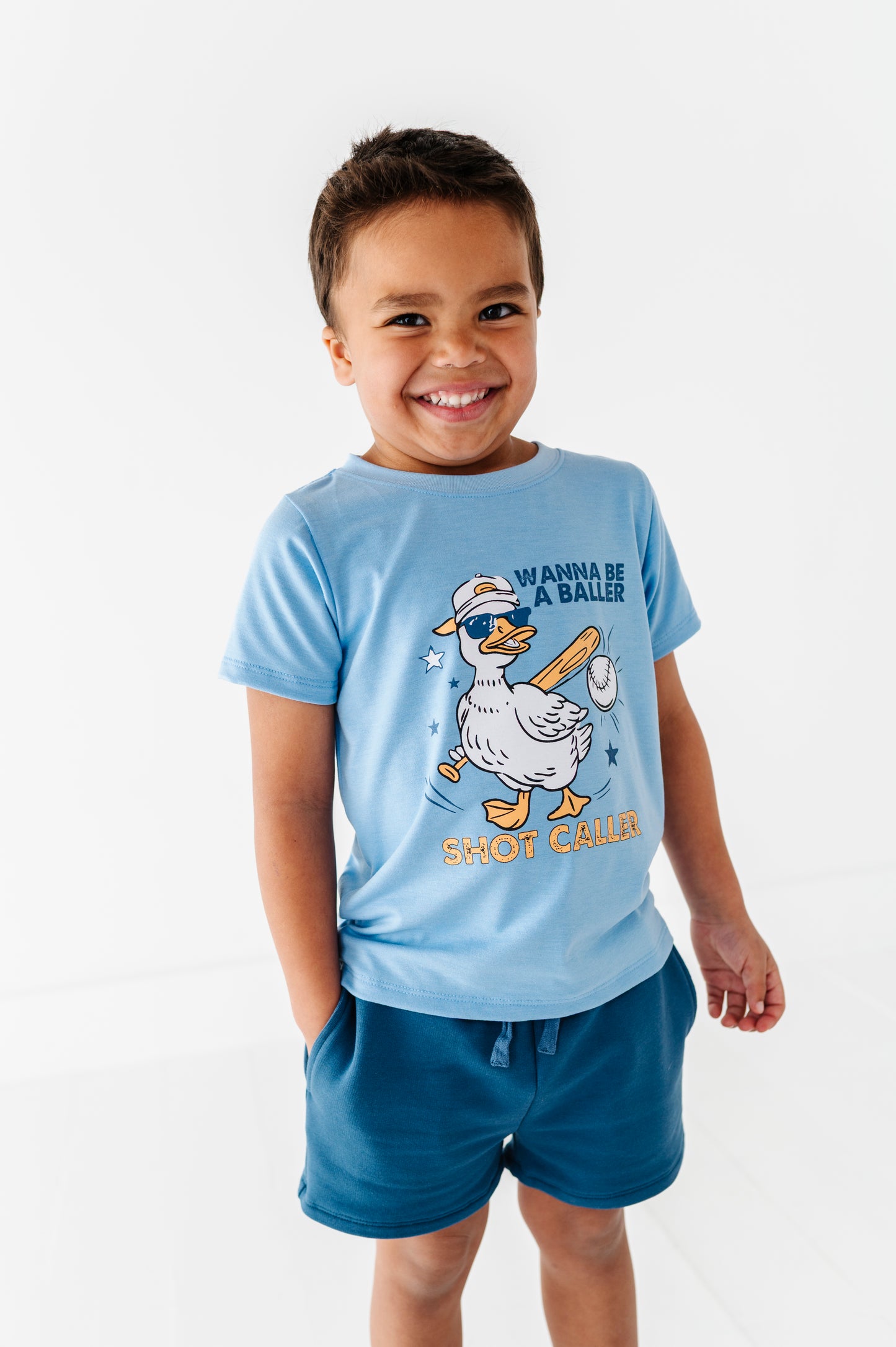 Child wearing a light blue baseball t-shirt with a cartoon duck graphic and text, standing against a white background.