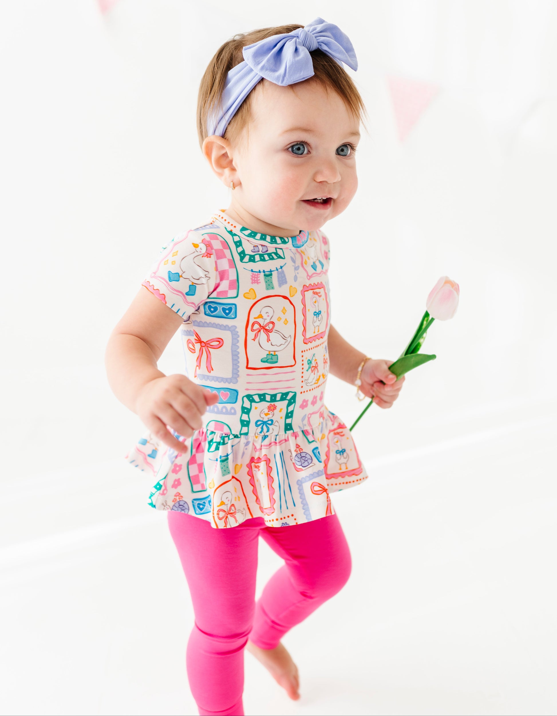 Child wearing a colorful bamboo/cotton blend peplum top with a pattern of bows and holding a pink flower on a white background