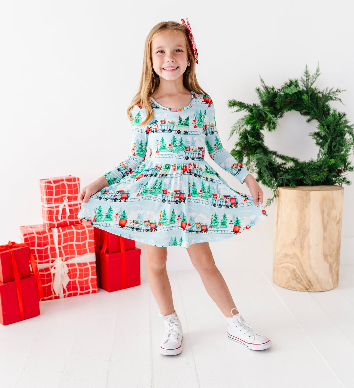 A young girl stands in a bright white room wearing a light blue long-sleeve bamboo dress with a Christmas tree and Santa pattern. She is holding out her skirt to show the full print, with red wrapped gift boxes on her left and a green wreath on a wooden pedestal on her right.