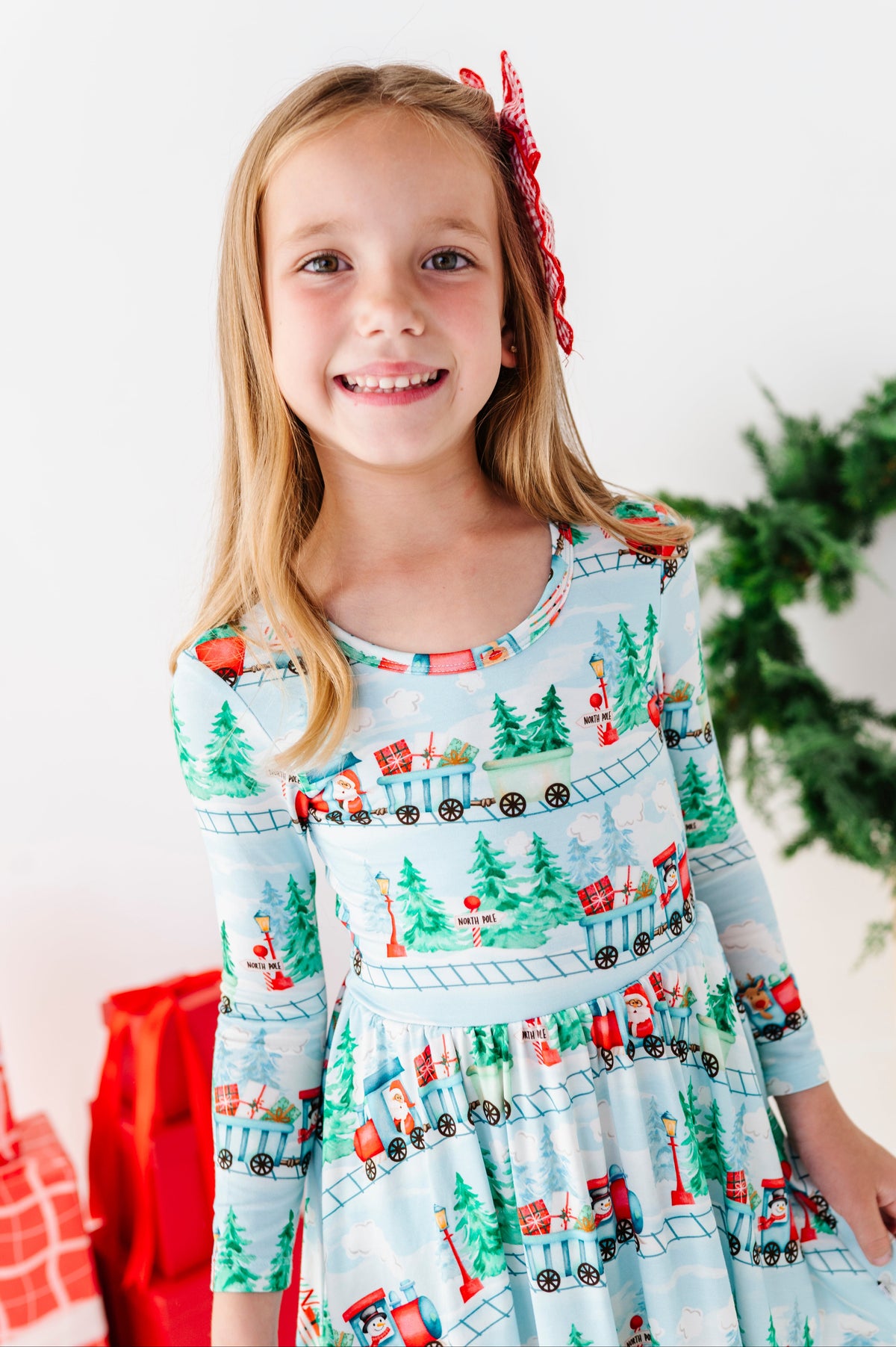 A young girl with blonde hair wearing a light blue long-sleeve bamboo dress with a Christmas train pattern, smiling at the camera.  
