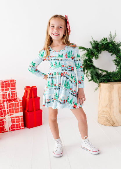 A young girl smiling and wearing a light blue long-sleeve bamboo dress with a Christmas train print, standing in a bright studio with wrapped red gifts, a green wreath, and a wooden stool.