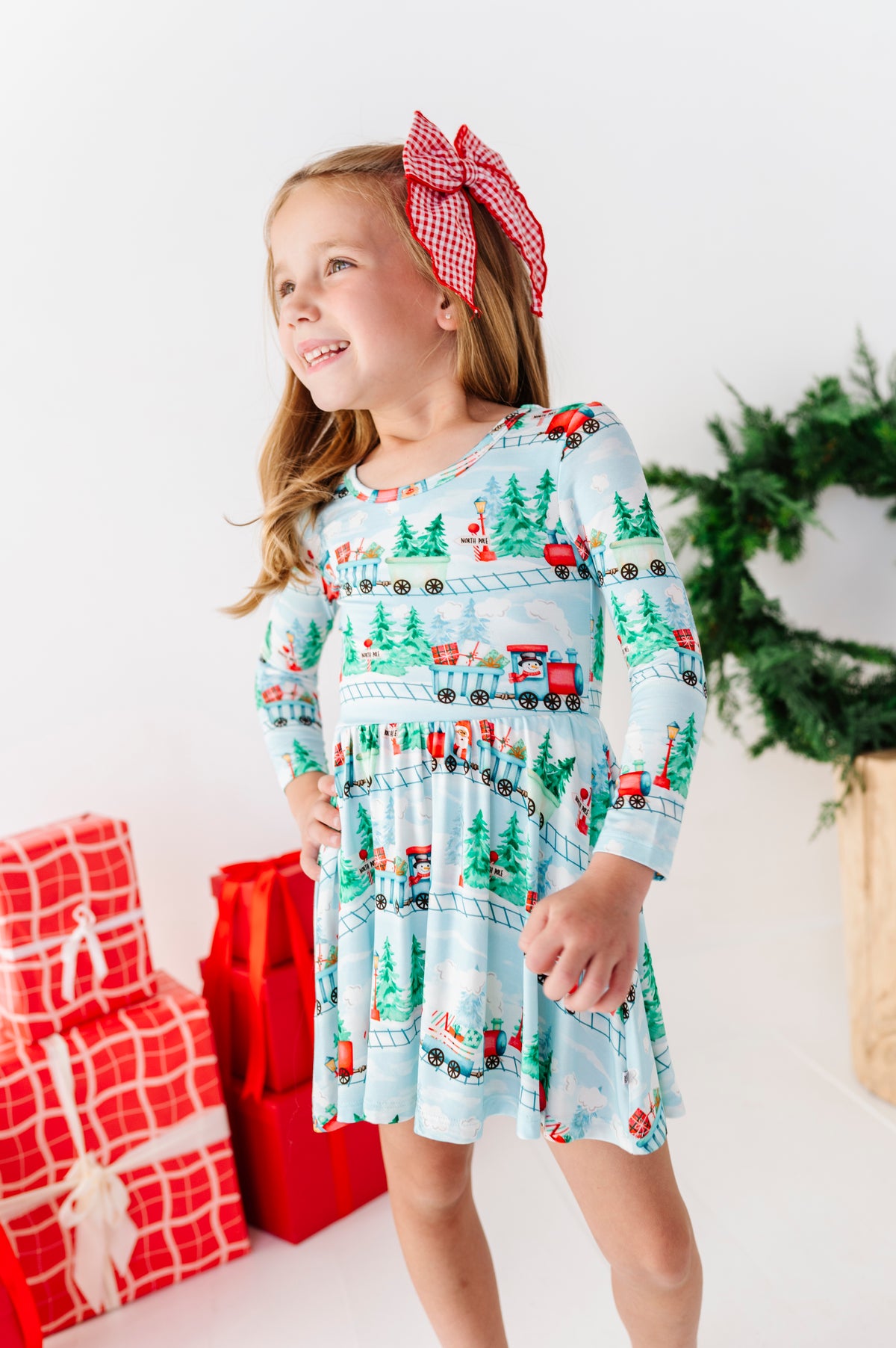 A young girl smiling and wearing a light blue long-sleeve bamboo dress with a Christmas train print, standing in a bright studio with wrapped red gifts.