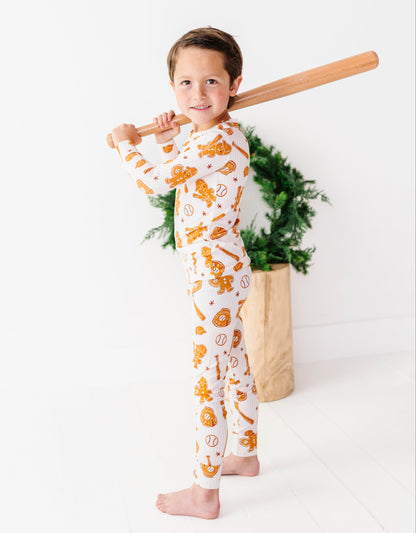 A young boy stands barefoot holding a wooden baseball bat over his shoulder, wearing a white long-sleeve pajama set with an orange cartoon character print. He is posed in front of a white wall with a potted green plant in the background.