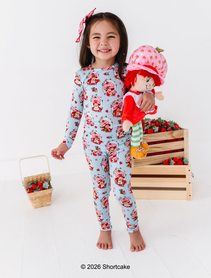 Child wearing a Strawberry Shortcake™ two-piece set holding a Strawberry Shortcake™ doll with a strawberry-themed background