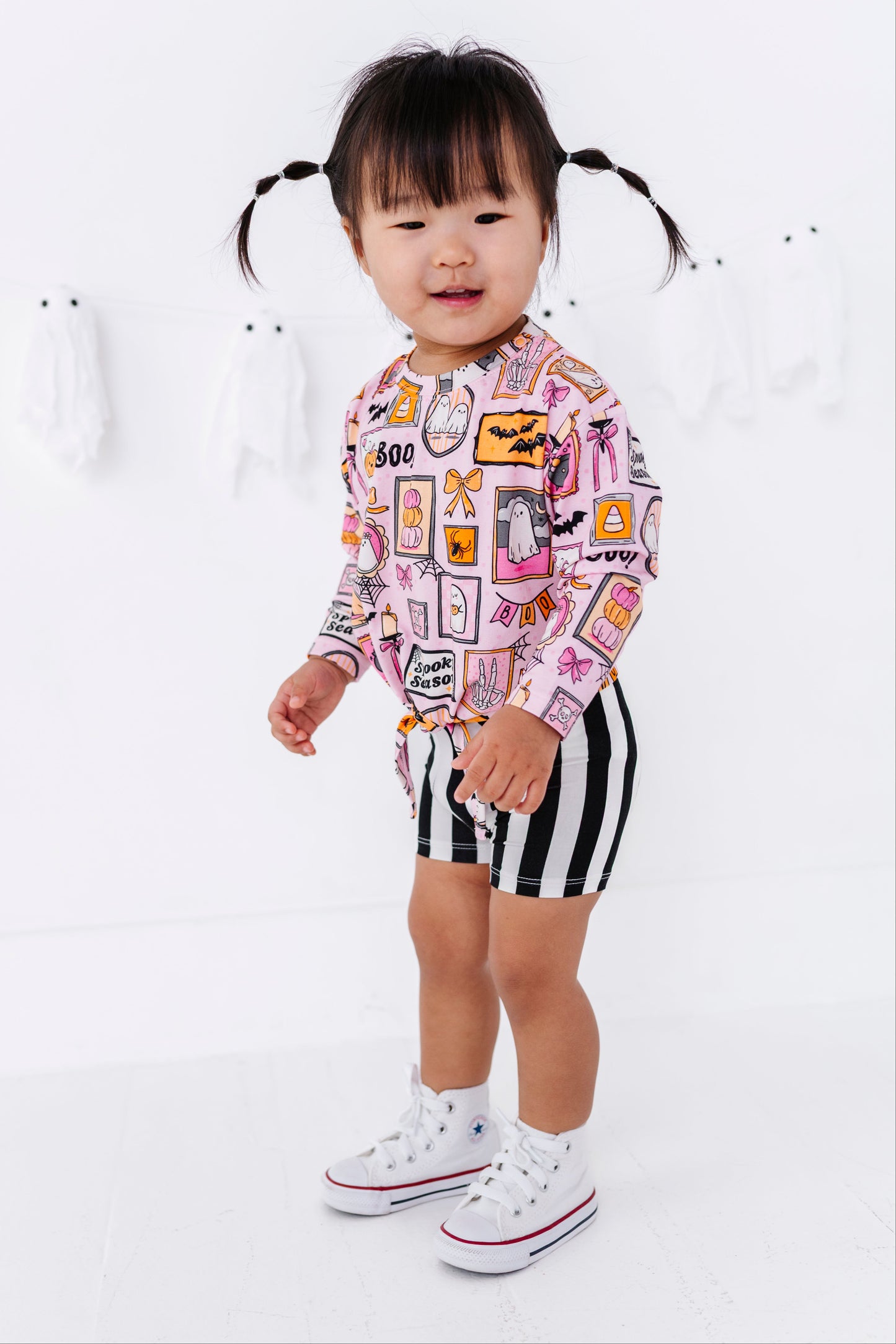 A toddler girl with black pigtails stands in a white studio setting, wearing a pink long-sleeve top covered in colorful graphic patches and black-and-white striped shorts, paired with white sneakers.