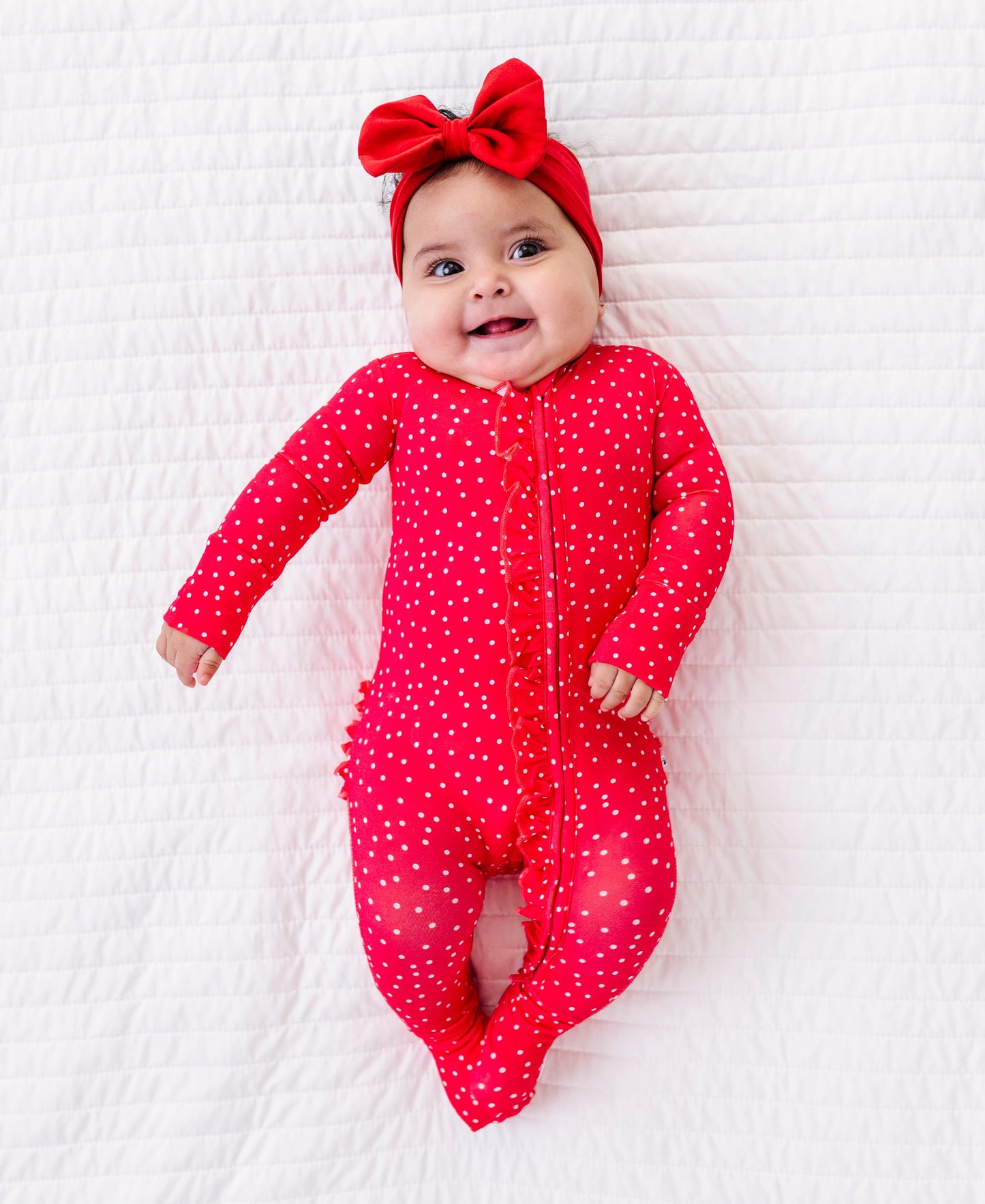 Baby wearing a red polka dot romper and headband on a white textured surface