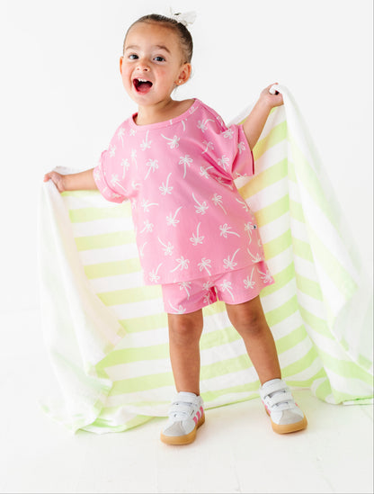 Child in a pink outfit with palm tree pattern standing on a yellow and white striped blanket.
