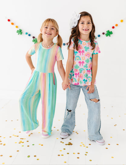 Two young girls holding hands, one in a colorful striped bamboo jumpsuit and the other in a bamboo cotton peplum top and ripped jeans, on a white background with confetti. 