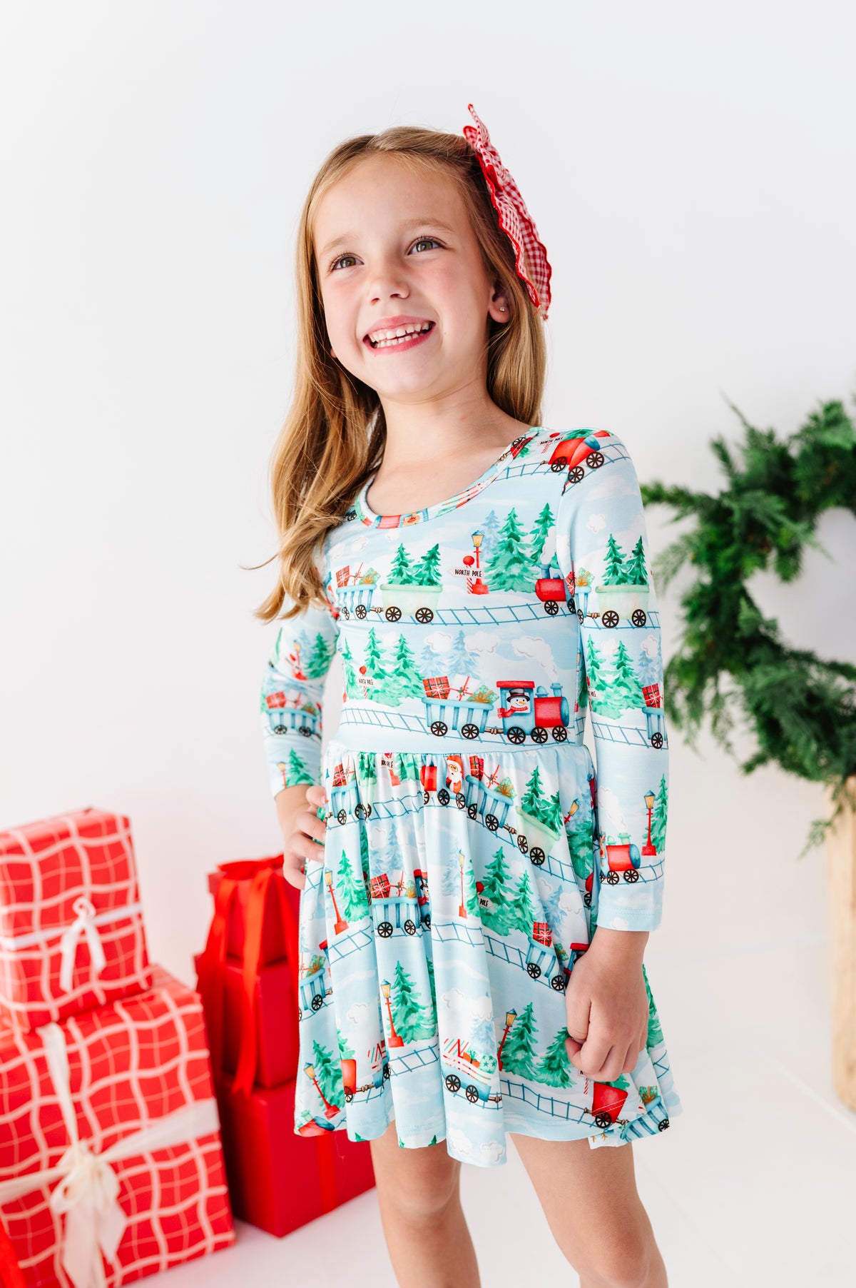 A young girl smiling and wearing a light blue long-sleeve bamboo dress with a Christmas train print, standing in a bright studio with wrapped red gifts, a green wreath, and a wooden stool.