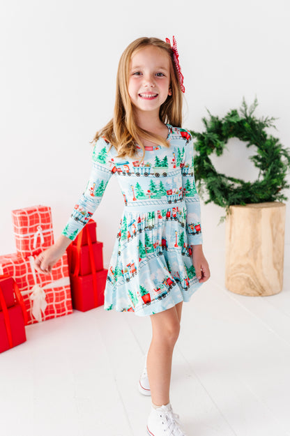 A young girl smiling and wearing a light blue long-sleeve bamboo dress with a Christmas train print, standing in a bright studio with wrapped red gifts, a green wreath, and a wooden stool.