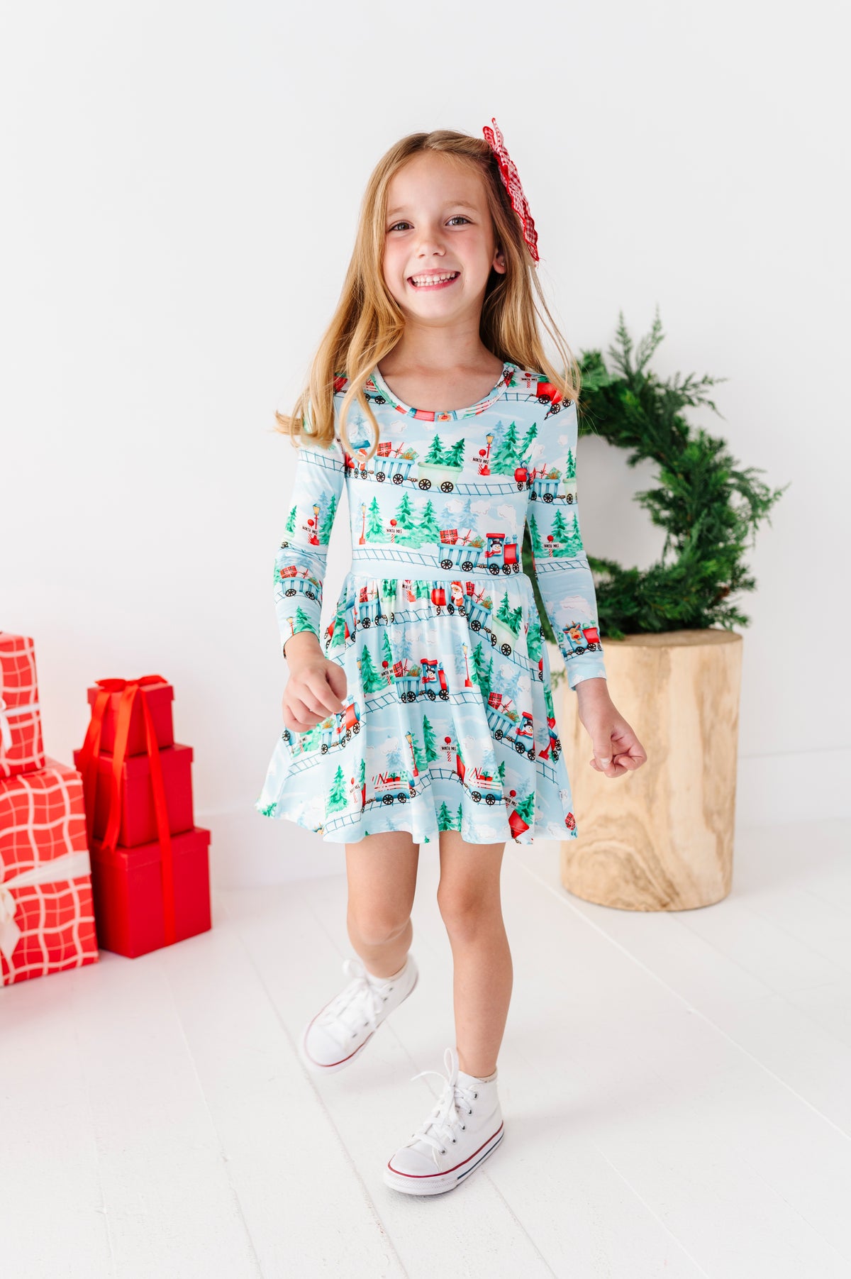 A young girl smiling and wearing a light blue long-sleeve bamboo dress with a Christmas train print, standing in a bright studio with wrapped red gifts, a green wreath, and a wooden stool.