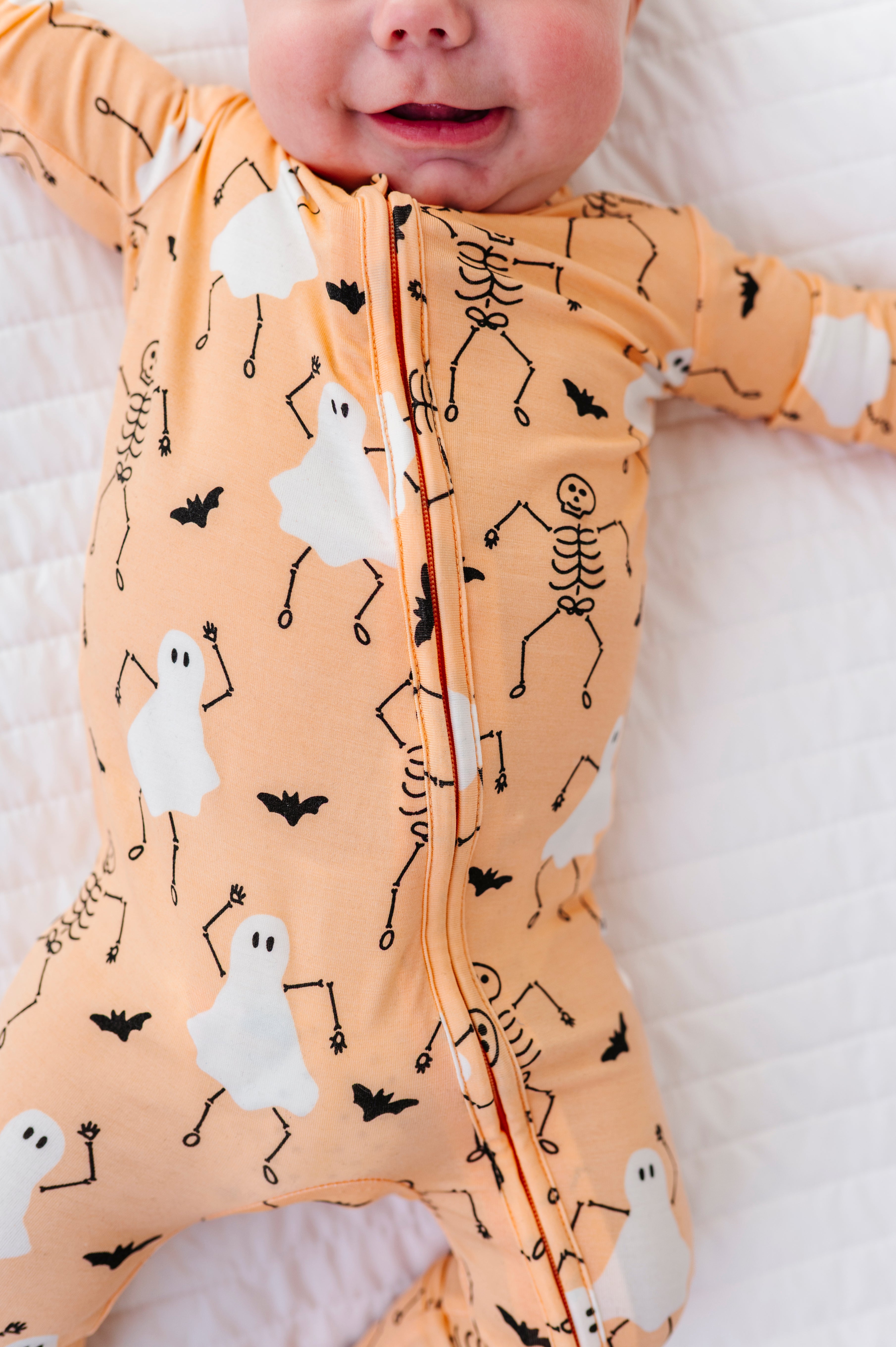 Baby wearing a Halloween-themed onesie with ghosts, skeletons, and bats on a white blanket.