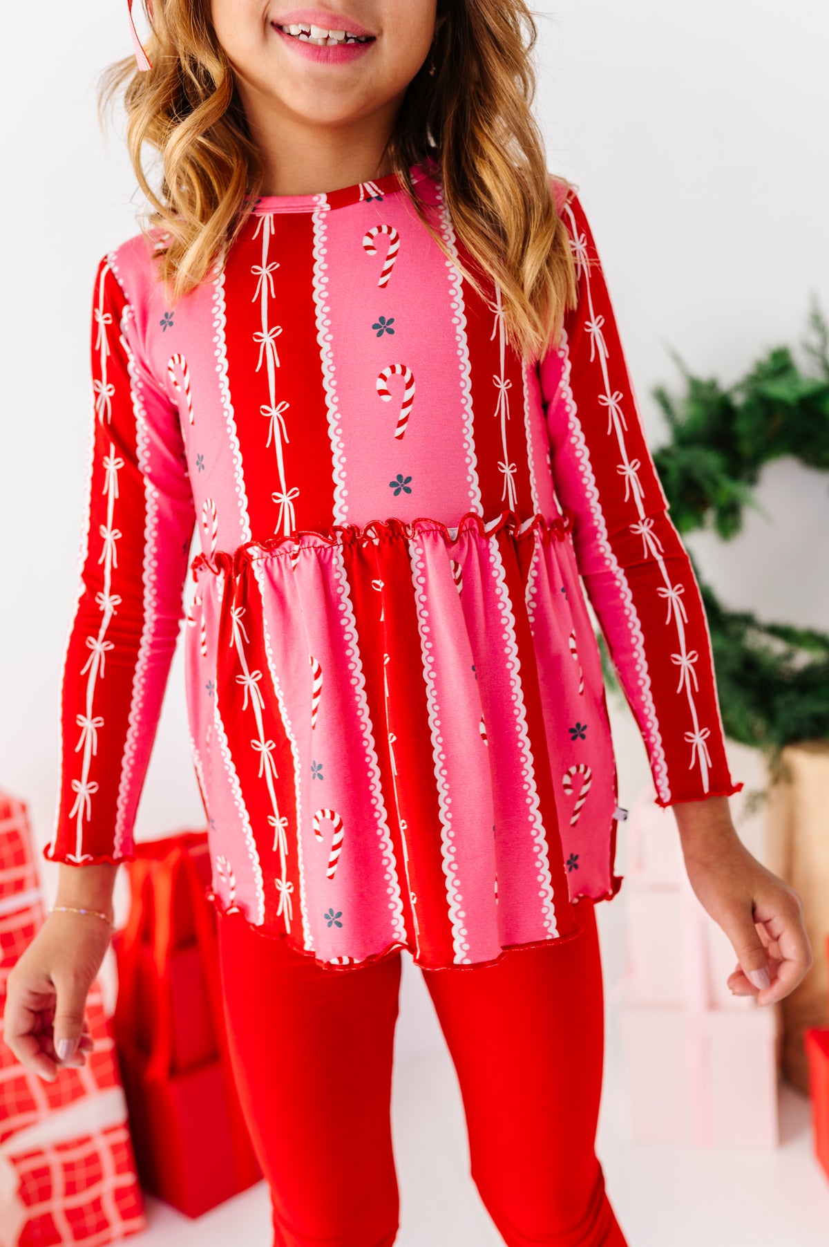 A child wearing a festive long-sleeve bamboo top with a red and pink holiday print of candy canes, snowflakes, and bows, paired with red leggings, set against a decorated Christmas background.