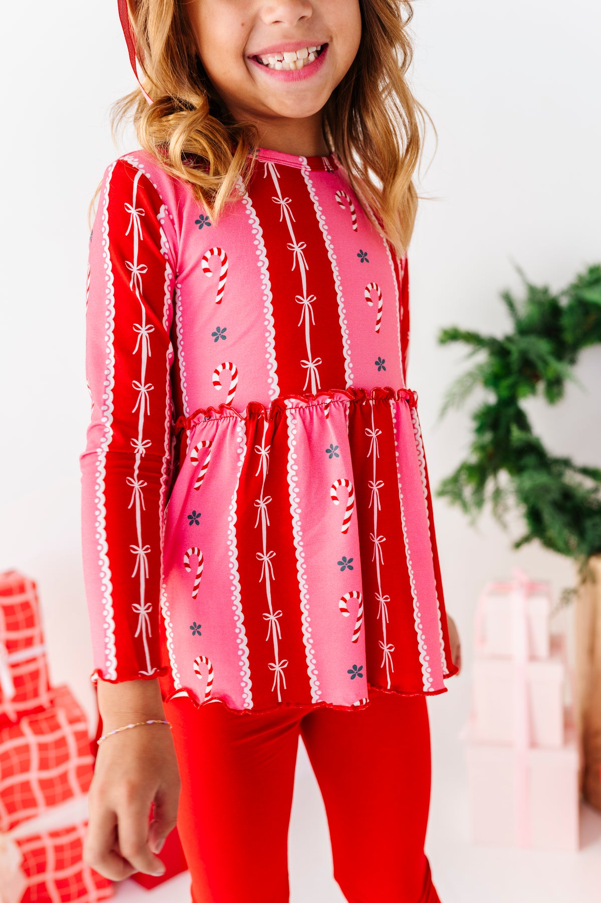 A young girl wearing a festive red and pink long-sleeve bamboo top with candy cane and bow patterns, paired with red leggings, standing in a holiday-themed setting with wrapped gifts and a plant in the background.