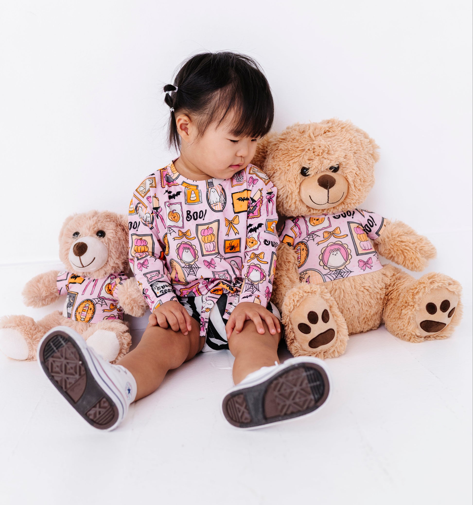 A young child wearing a pink long-sleeve top with a colorful cartoon print sits on a white floor between two teddy bears, one small and one large.