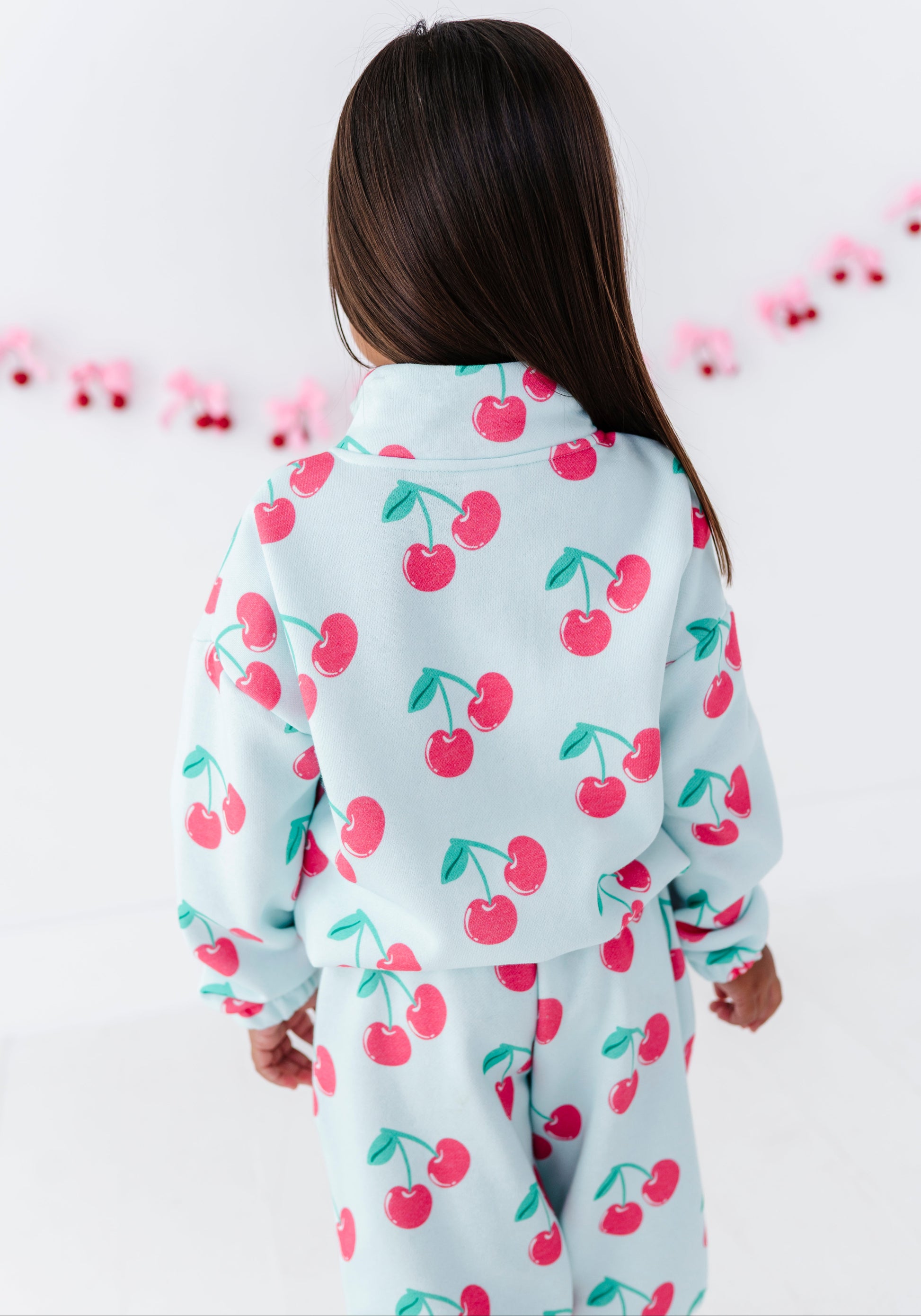 A child viewed from behind wearing a light blue two-piece pajama set with a red cherry print, set against a white background with a pink garland.