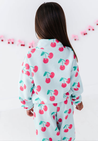 A child viewed from behind wearing a light blue two-piece pajama set with a red cherry print, set against a white background with a pink garland.