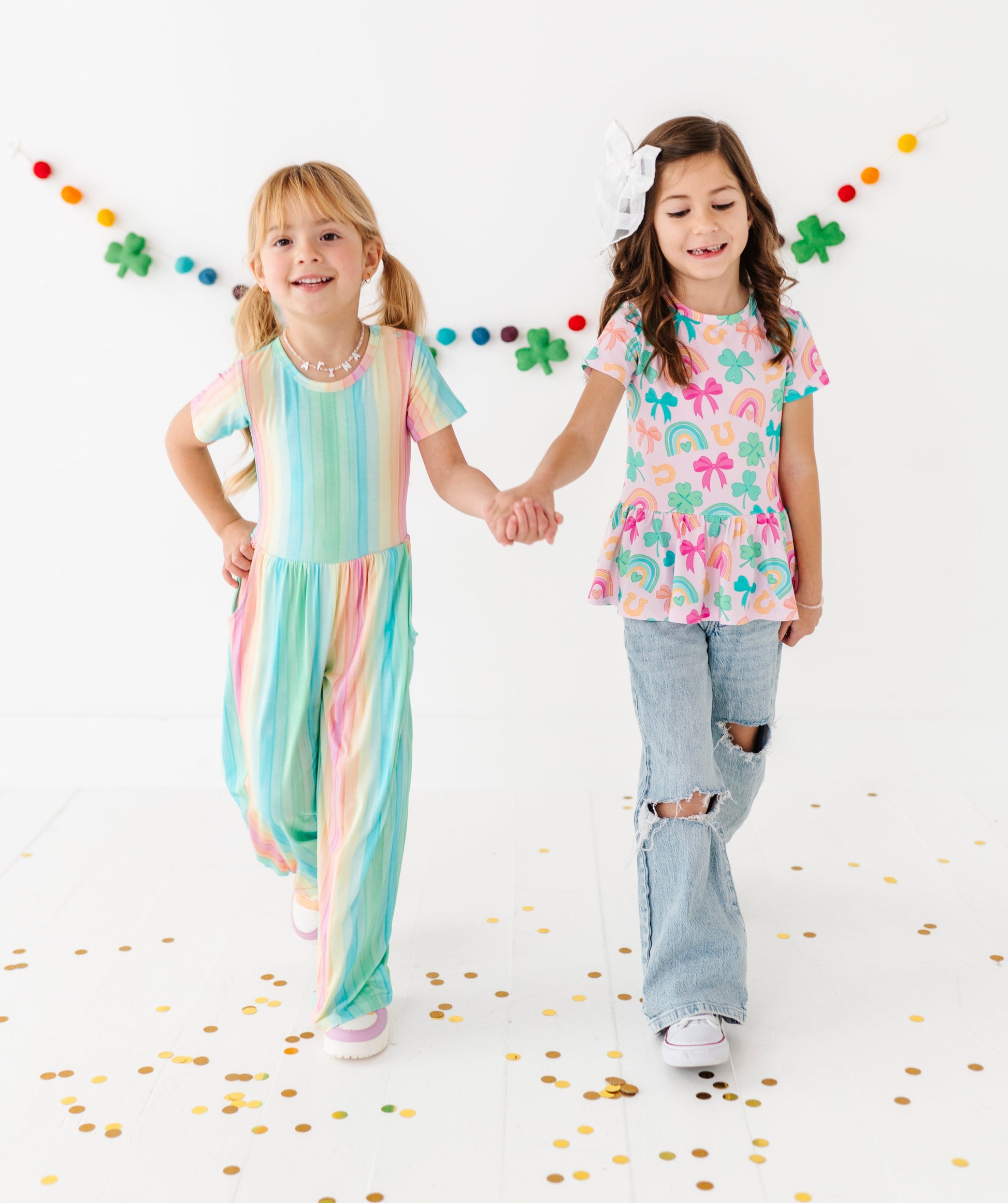 Two young girls holding hands on a white background with colorful confetti and decorations. The girls are wearing bamboo St. Patrick's Day outfits