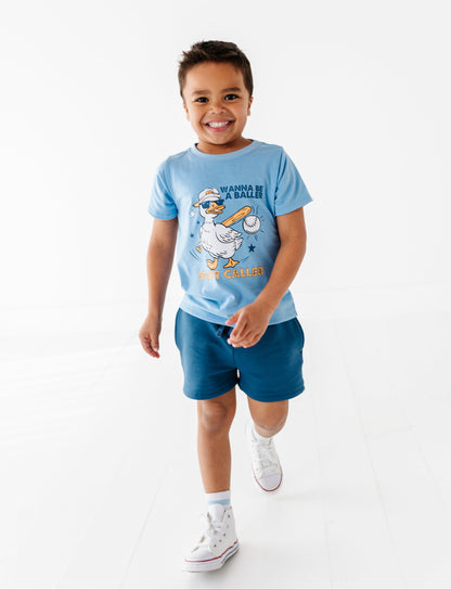 Child wearing a blue baseball-themed t-shirt with a cartoon character and text, standing on a white background