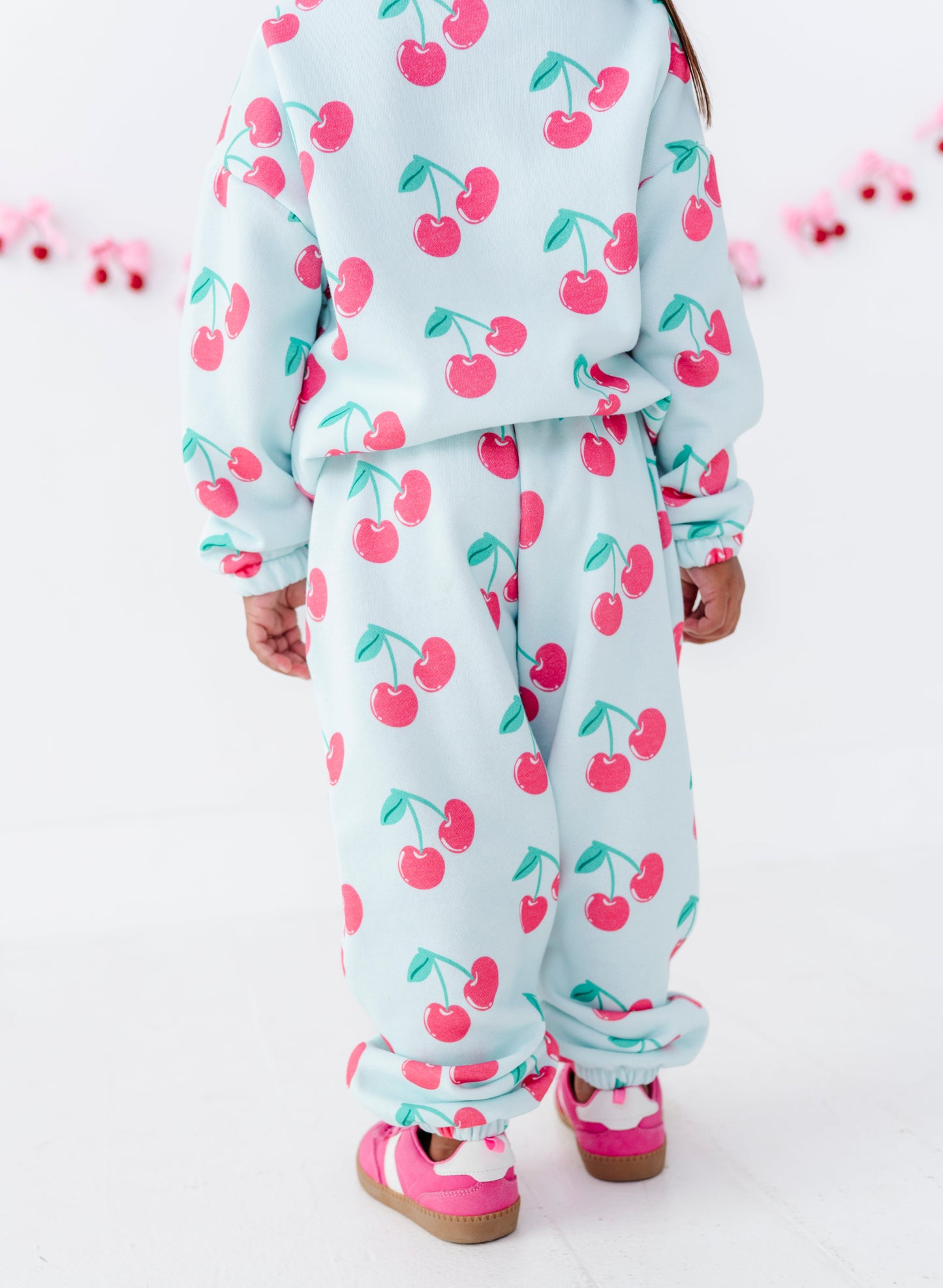 A child wearing a matching light blue cherry-print sweatshirt and jogger pants set, paired with pink sneakers, shown from the back against a white studio background.