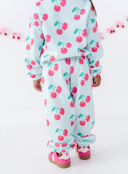 A child wearing a matching light blue cherry-print sweatshirt and jogger pants set, paired with pink sneakers, shown from the back against a white studio background.