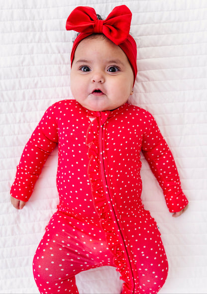 Baby in a red polka dot outfit with a large red bow on a white textured surface