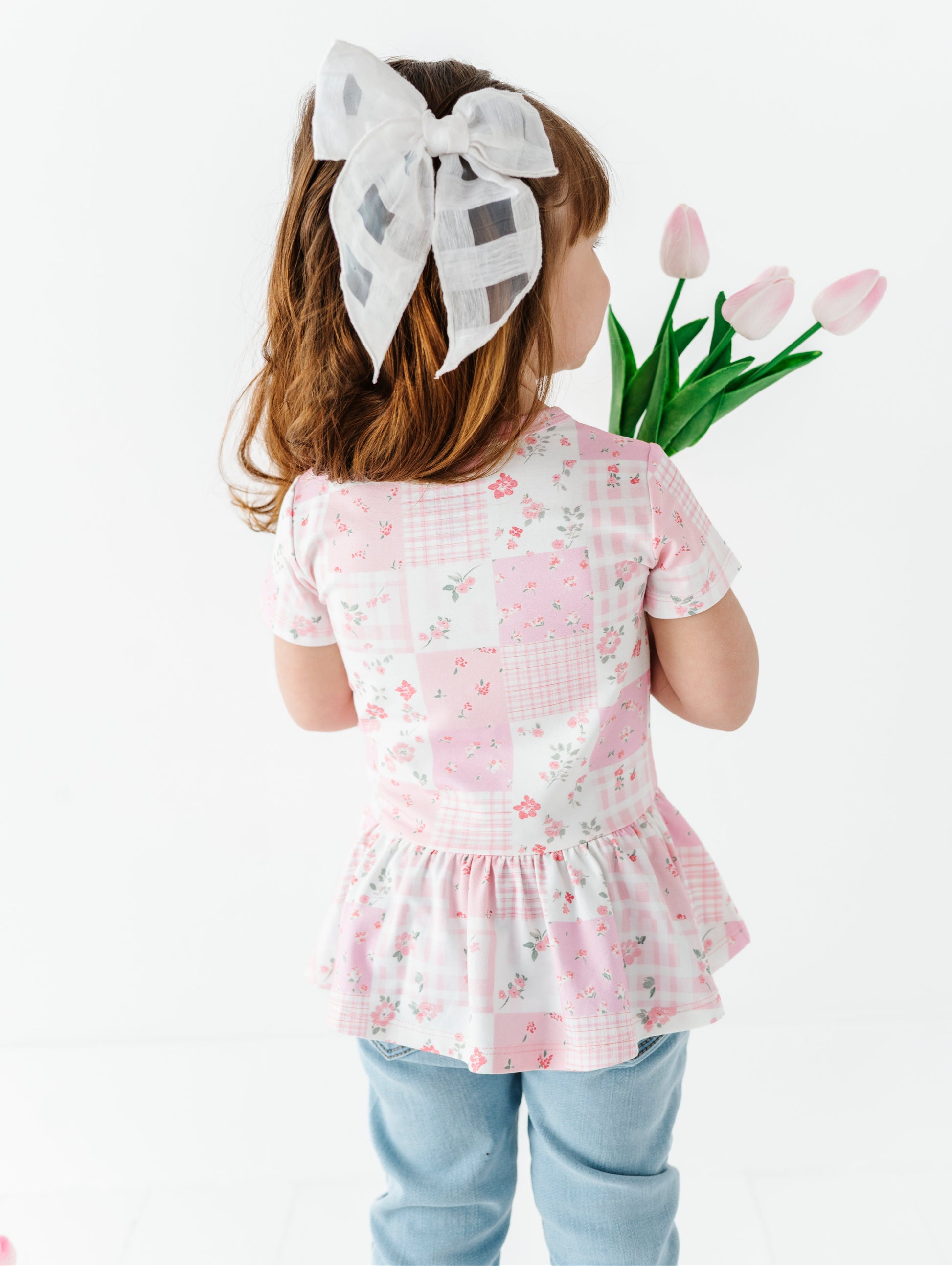 Child wearing a pink floral bamboo-cotton peplum blouse with a white bow, holding tulips against a white background
