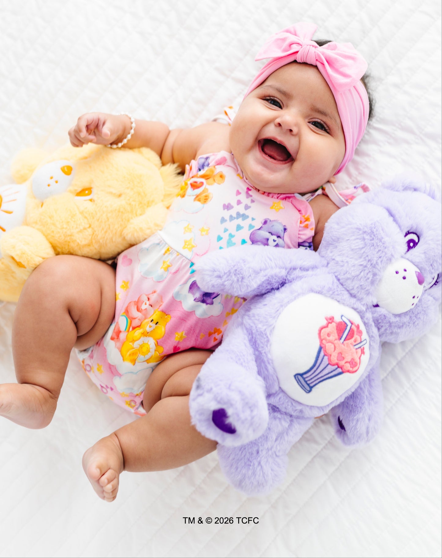 Baby holding a purple Care Bears™ toy with a smile on a white background