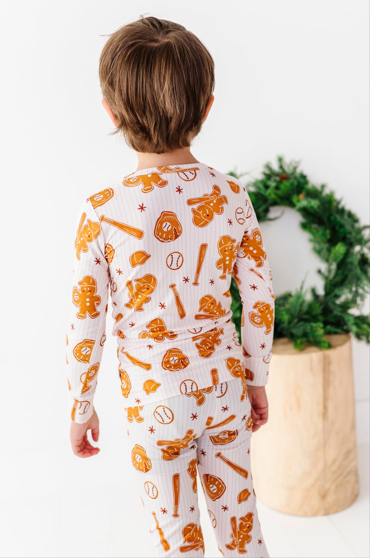 A child viewed from behind wearing a white long-sleeve pajama set with an orange baseball-themed print, standing in a bright indoor setting next to a green wreath in a wooden pot.