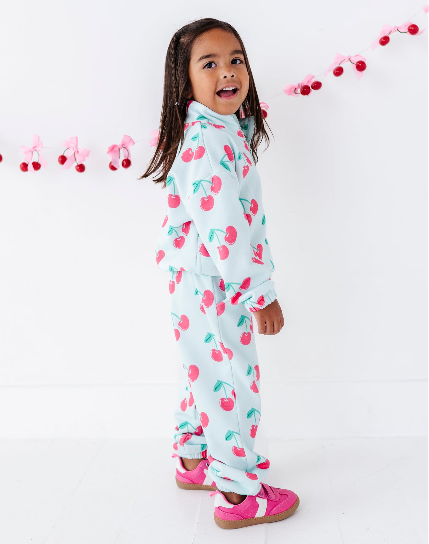A young girl with dark hair is wearing a light blue two-piece pajama set with a pink and green cherry print, standing against a white wall with a pink cherry garland.