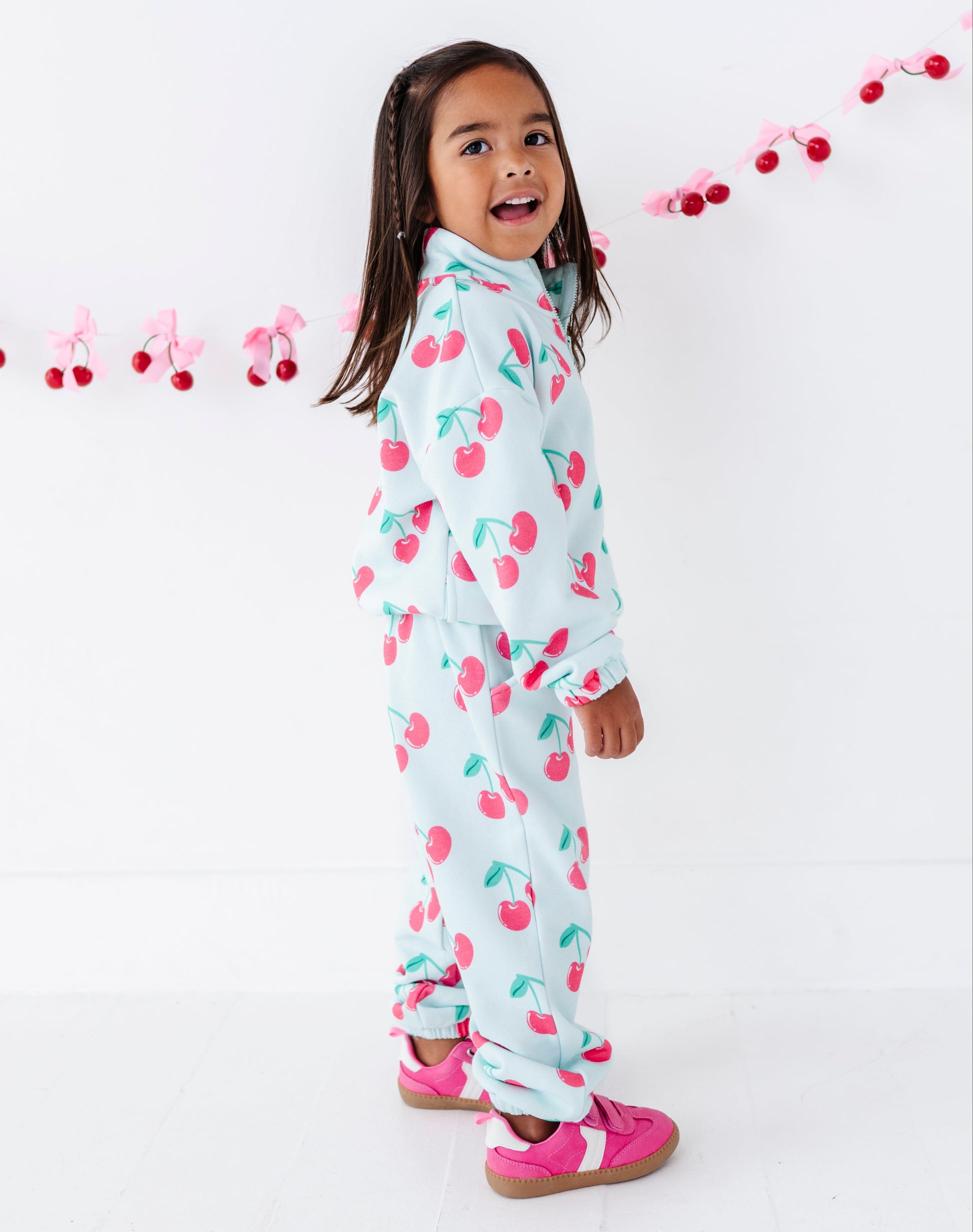 A young girl with dark hair is wearing a light blue two-piece pajama set with a pink and green cherry print, standing against a white wall with a pink cherry garland.