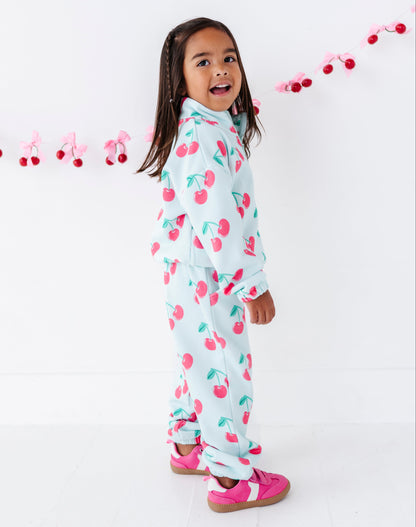 A young girl with dark hair is wearing a light blue two-piece pajama set with a pink and green cherry print, standing against a white wall with a pink cherry garland.