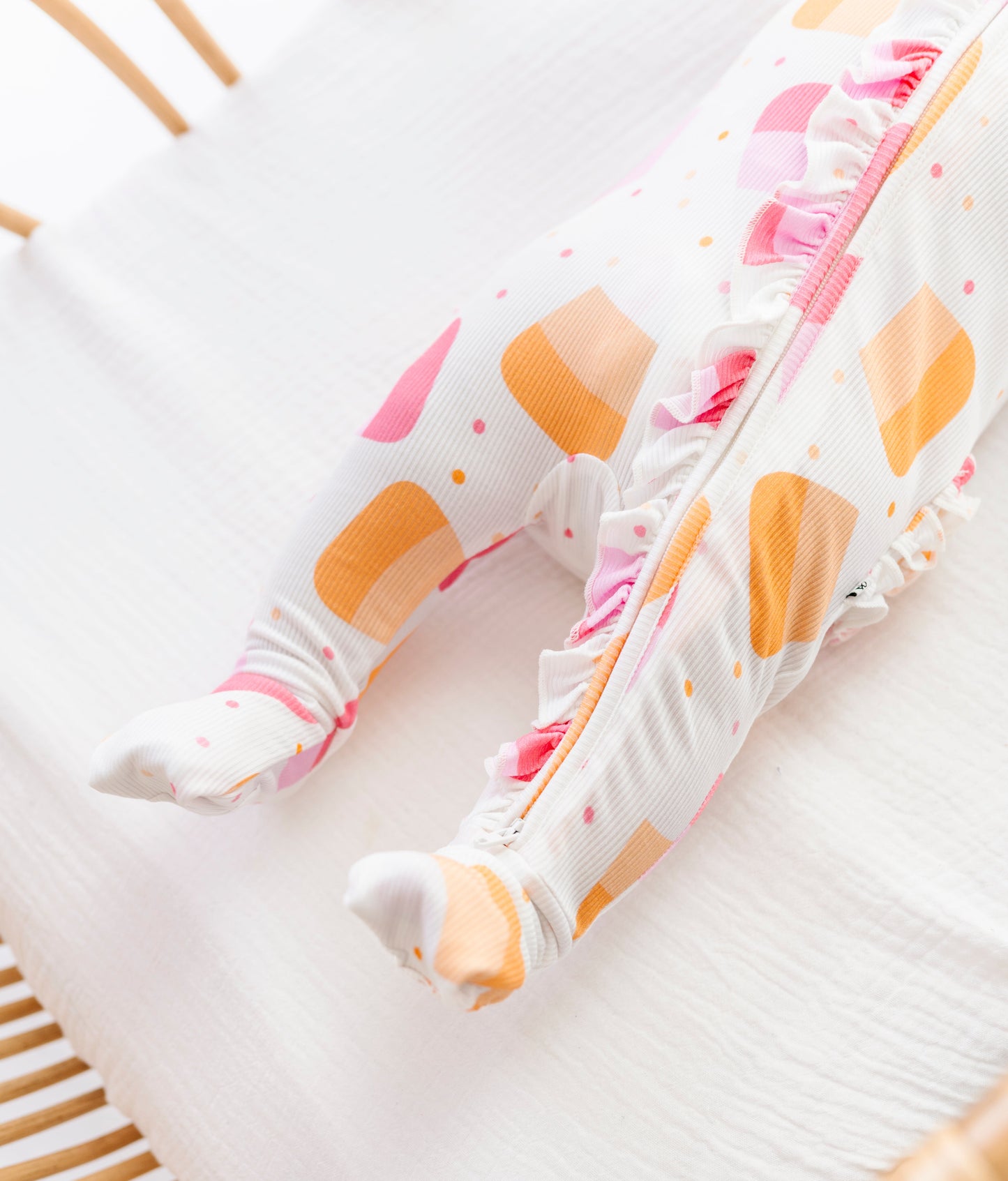 A close-up of a baby's feet wearing white footed pajamas with orange and pink geometric patterns, resting on a white muslin blanket in a wooden crib.